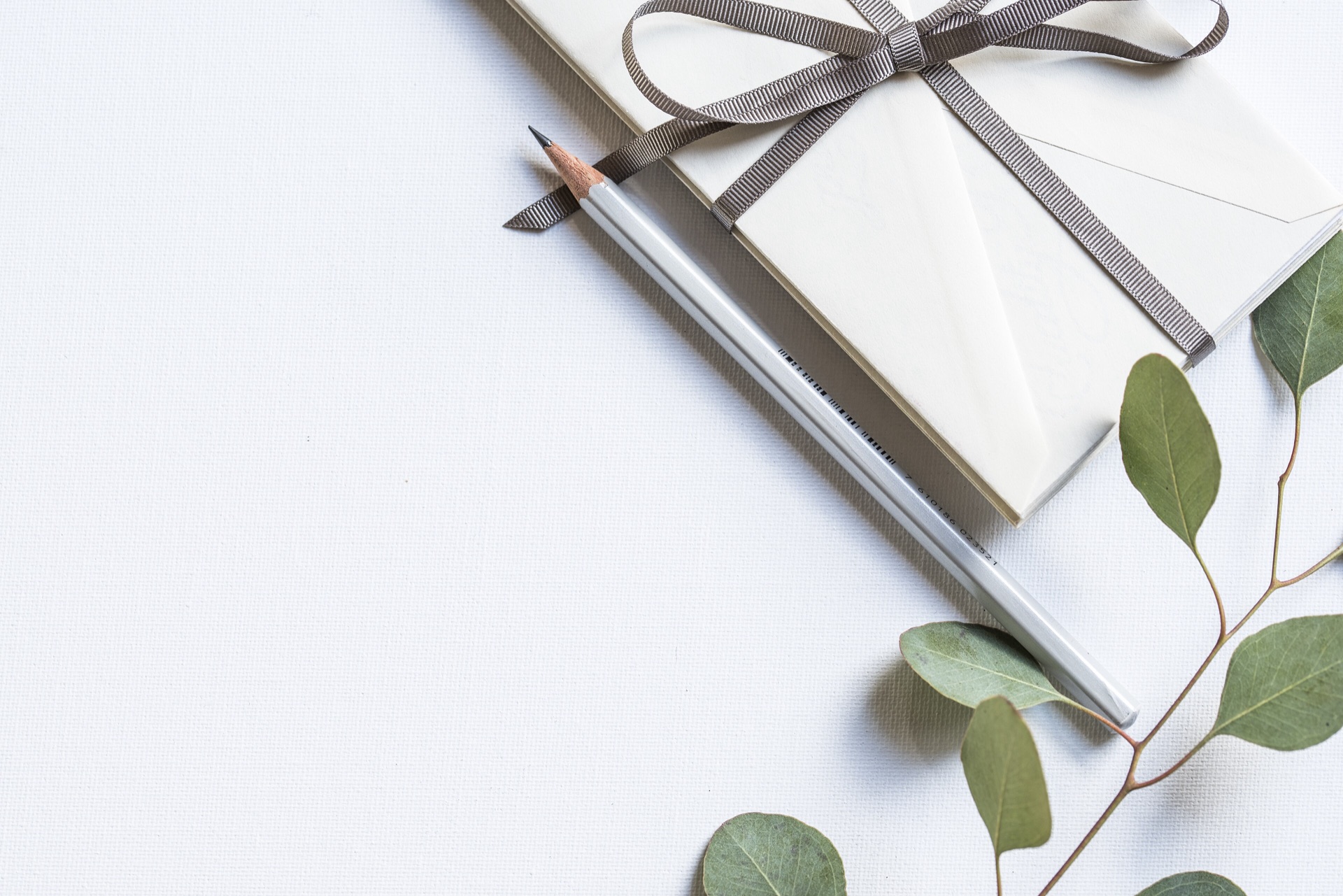 Feuille de papier blanc texturé, enveloppes crème nouées d'un ruban, crayon et branche d'eucalyptus