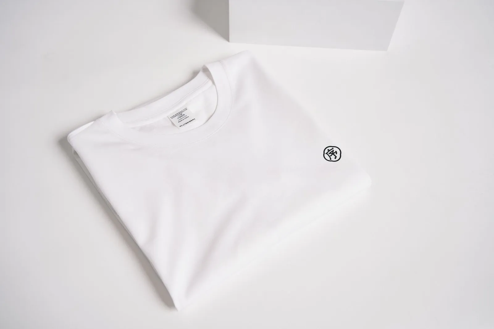 T-shirt blanc plié sur fond blanc, avec un petit motif discret sur la poitrine
